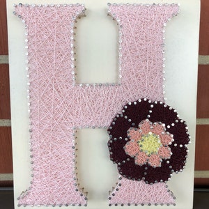 May include: A pink string art letter H with a burgundy flower with a pink and yellow center.