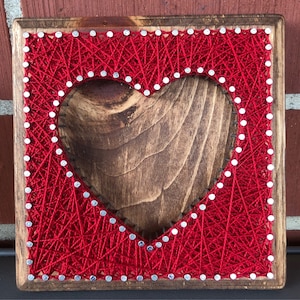 May include: A square wooden string art piece with a heart-shaped cutout. The heart is outlined with small silver nails and filled with red string. The wooden frame has a natural wood grain finish.