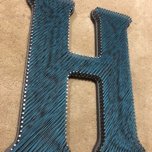 May include: A large, teal-colored letter "H" made with string art. The letter is outlined in gray and has small silver nails along the edges. The string art creates a textured effect, suitable for home decor.