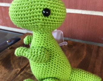May include: A green crocheted dinosaur toy with black eyes. The dinosaur is sitting on a wooden surface.