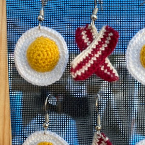 May include: Crocheted earrings shaped like fried eggs and bacon. The eggs are yellow and white, and the bacon is red and white.