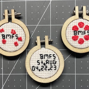 May include: Three small wooden embroidery hoops with white fabric and red embroidery. The hoops are hanging from a screw. The fabric in the first hoop has a red flower design and the text "BMFS". The fabric in the second hoop has the text "BMFS 5+AUG 04.22.23". The fabric in the third hoop has a red flower design and the text "BMFS".
