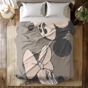 Peut inclure: Une couverture au design pixelisé représentant le visage et les mains d'un personnage de dessin animé dans des tons de gris, de noir et de beige. La couverture est posée sur un lit avec des oreillers blancs.