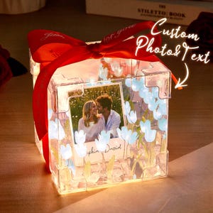 Può includere: Una scatola regalo in acrilico trasparente illuminata, con un nastro rosso e una foto di una coppia. La scatola è decorata con motivi floreali blu e bianchi e il testo "Custom Photo & Text". La foto ha i nomi "Sophia David".
