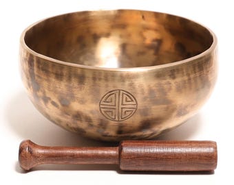 May include: A metallic bronze-colored singing bowl with a hammered texture and a circular geometric symbol etched on the front. The bowl is accompanied by a straight, dark brown wooden mallet with a cylindrical handle and a rounded end.