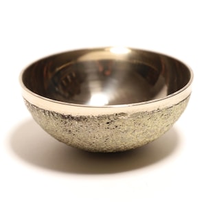 Op de afbeelding: Een kleine, ronde kom met een getextureerde, lichtgouden buitenkant en een gladde, zilverkleurige binnenkant. De kom heeft een eenvoudig, elegant ontwerp, perfect voor het serveren van kleine porties of als decoratief accent. De kom heeft een diameter van ongeveer 7.6 cm.