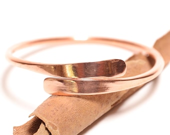 Könnte beinhalten: Ein poliertes, kupferfarbenes Metall-Armband mit offenem Bypass-Design und überlappenden Enden, das auf einer einzelnen, strukturierten, hellbraunen Zimtstange vor einem schlichten weißen Hintergrund liegt.
