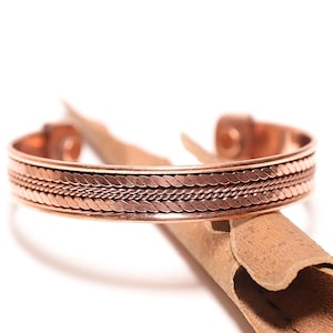 Peut inclure: Un bracelet manchette en cuivre avec un motif tressé. Le bracelet est d'une couleur brun rougeâtre chaud et semble être en métal. Le bracelet repose sur un morceau de matériau brun.