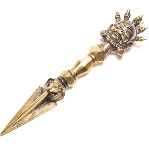 May include: A brass vajra, a ritual object with a detailed design. The handle features a detailed face and skull design, with a pointed blade at the end. The object is golden in color and sits against a white background.