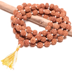 May include: A mala necklace featuring numerous round, textured, brown rudraksha beads strung on a white cord. The beads are arranged in multiple loops and finished with a small, bright yellow tassel at the end of the strand, creating a circular design.