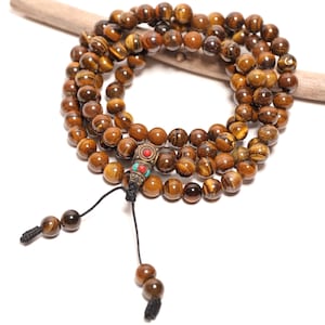 May include: A multi-strand necklace composed of polished brown and gold tiger eye beads. The necklace features a central metal spacer bead inlaid with small red and turquoise stones, finished with black cord ends and two dangling tiger eye beads.