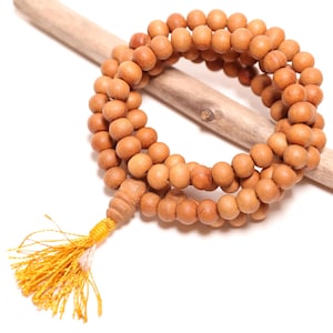 May include: A strand of round, light brown wooden beads arranged in a circular loop, featuring a single larger bead and a bright yellow decorative tassel hanging from the bottom, resting across a smooth, light-colored piece of driftwood.