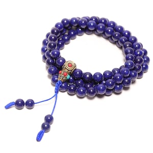 May include: A multi-strand bracelet composed of deep blue spherical beads. The piece features a central metallic bead with intricate engravings and small inlaid red and turquoise accents, finished with two blue cord tassels ending in blue beads.
