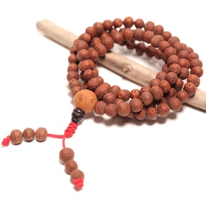 May include: A strand of brown, textured, spherical wooden beads arranged in a circular loop. The beads are strung on a red cord, featuring a larger, lighter-colored bead at the base, followed by a dark bead and two dangling ends with smaller beads.