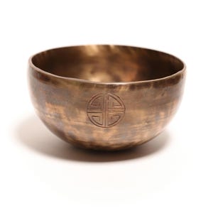 May include: A round, metallic singing bowl with a bronze-colored finish and a mottled, darkened patina. The exterior surface features a centered, circular engraved symbol consisting of four symmetrical quadrants enclosed within a thin ring.