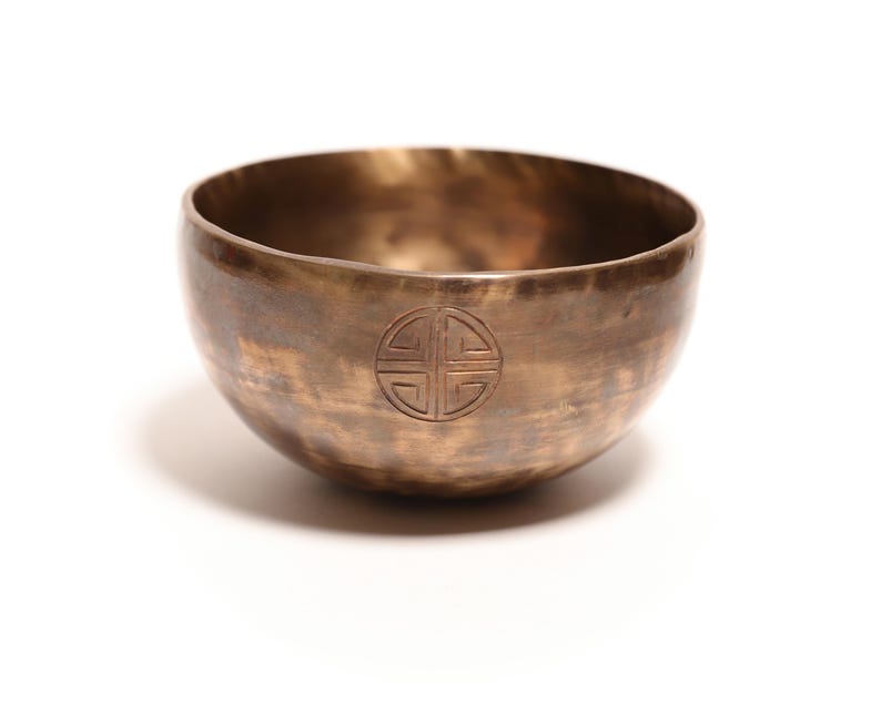 May include: A round, metallic singing bowl with a bronze-colored finish and a mottled, darkened patina. The exterior surface features a centered, circular engraved symbol consisting of four symmetrical quadrants enclosed within a thin ring.