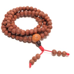 May include: A strand of brown, textured, spherical wooden beads arranged in a circular loop. A larger, lighter-colored bead is positioned near the bottom, followed by a dark brown bead, red cord, and two dangling ends with three small beads each.