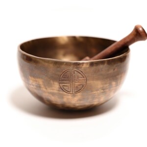 May include: A round, metallic bronze singing bowl with a hammered texture and a circular engraved symbol on the front. A dark brown wooden mallet rests inside the bowl, angled against the inner rim. The bowl has a polished, reflective finish.