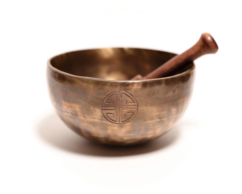 May include: A round, metallic bronze singing bowl with a hammered texture and a circular engraved symbol on the front. A dark brown wooden mallet rests inside the bowl, angled against the inner rim. The bowl has a polished, reflective finish.