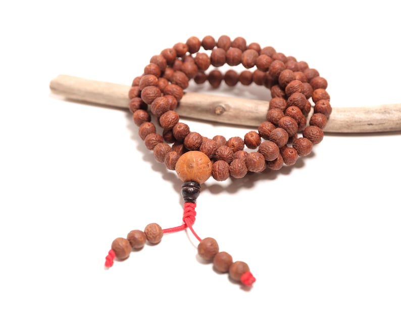 May include: A prayer bead necklace featuring numerous small, round, textured brown wooden beads arranged in a circular loop. A larger, lighter brown bead is positioned at the bottom, followed by a dark bead and red cord ends with additional beads.