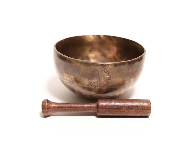 May include: A metallic bronze singing bowl with a dark, mottled patina and a circular engraved symbol on the front. The bowl is accompanied by a straight, cylindrical wooden mallet with a smooth, dark brown grain finish and rounded ends.