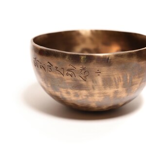 May include: A round, metallic singing bowl with a warm bronze finish and a hammered texture. The exterior surface features an engraved inscription in Tibetan script along the upper rim, showcasing a dark, weathered patina with subtle color variations.