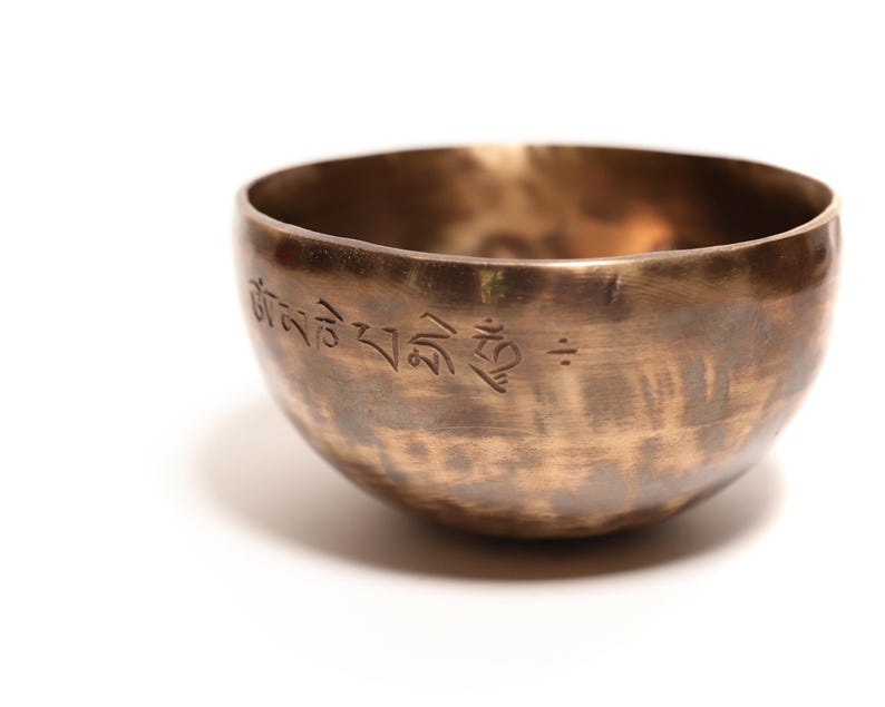 May include: A round, metallic singing bowl with a warm bronze finish and a hammered texture. The exterior surface features an engraved inscription in Tibetan script along the upper rim, showcasing a dark, weathered patina with subtle color variations.
