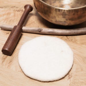 May include: A metallic bronze singing bowl with a dark wooden mallet, a thin wooden stick, and a circular white felt pad. The items are arranged together, featuring a polished metal finish on the bowl and a soft, textured surface on the felt pad.