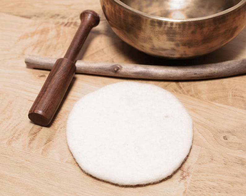 May include: A metallic bronze singing bowl with a dark wooden mallet, a thin wooden stick, and a circular white felt pad. The items are arranged together, featuring a polished metal finish on the bowl and a soft, textured surface on the felt pad.