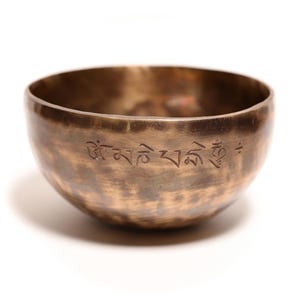 May include: A round, metallic singing bowl with a dark, burnished bronze finish and a hammered texture. The exterior surface features an engraved inscription in Tibetan script, encircling the upper portion of the bowl's curved, open-top body.