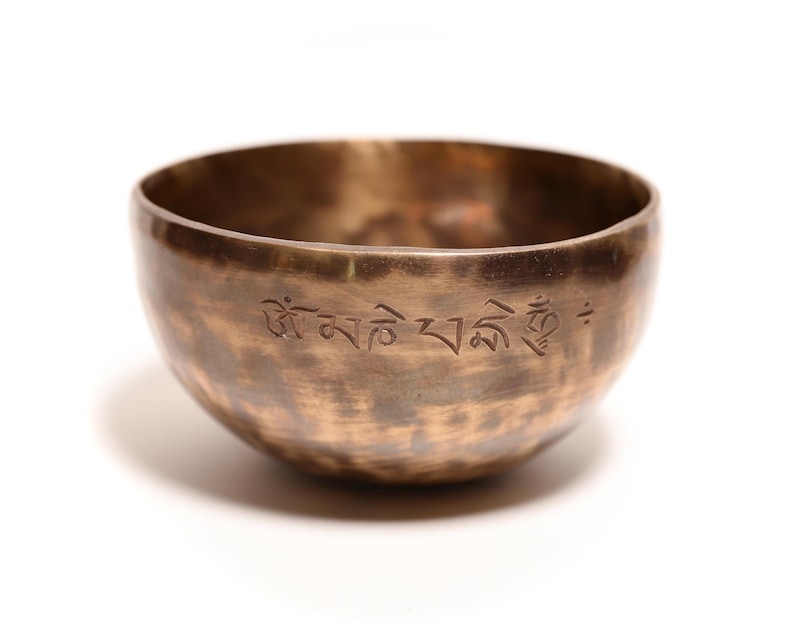 May include: A round, metallic singing bowl with a dark, burnished bronze finish and a hammered texture. The exterior surface features an engraved inscription in Tibetan script, encircling the upper portion of the bowl's curved, open-top body.