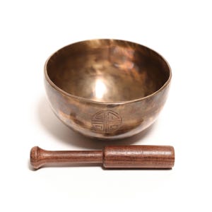 May include: A metallic bronze-colored singing bowl with a smooth, rounded interior and a dark, engraved circular symbol on the exterior. A straight, cylindrical wooden mallet with a slightly flared end is positioned horizontally in front of the bowl.