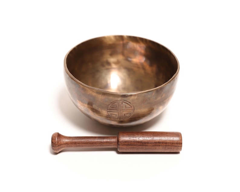 May include: A metallic bronze-colored singing bowl with a smooth, rounded interior and a dark, engraved circular symbol on the exterior. A straight, cylindrical wooden mallet with a slightly flared end is positioned horizontally in front of the bowl.