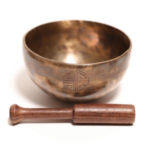 May include: A metallic bronze singing bowl with a circular engraved symbol on the front, accompanied by a dark brown wooden mallet. The bowl has a smooth, polished interior and a slightly textured, weathered exterior finish with visible dark patina.