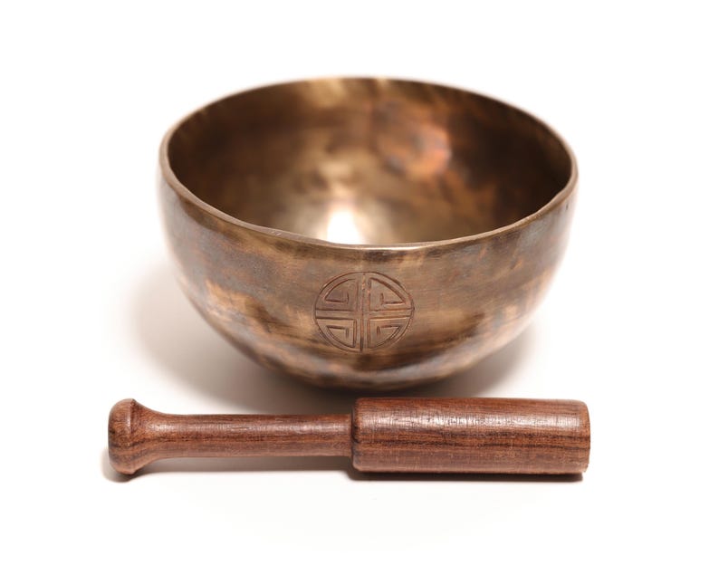 May include: A metallic bronze singing bowl with a circular engraved symbol on the front, accompanied by a dark brown wooden mallet. The bowl has a smooth, polished interior and a slightly textured, weathered exterior finish with visible dark patina.