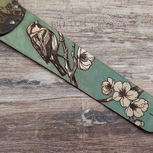 May include: A teal guitar strap with a detailed illustration of a bird perched on a flowering branch. The bird and flowers are in a contrasting light color, creating a striking visual against the teal background.