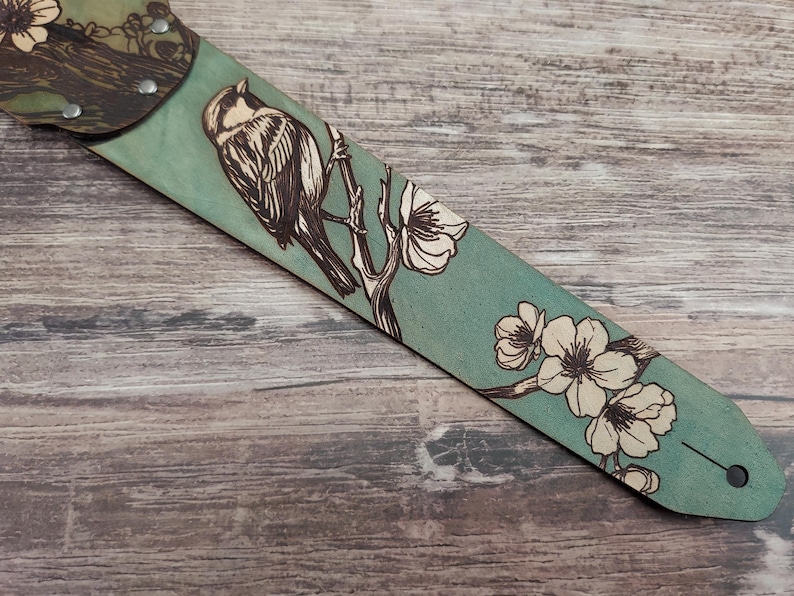 May include: A teal guitar strap with a detailed illustration of a bird perched on a flowering branch. The bird and flowers are in a contrasting light color, creating a striking visual against the teal background.
