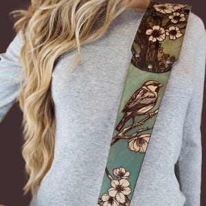 May include: A leather guitar strap with a hand-tooled design featuring a bird perched on a flowering branch. The strap has a teal background with brown and cream-colored details. The top portion has a tree and flower design.