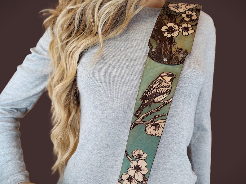 May include: A leather guitar strap with a hand-tooled design featuring a bird perched on a flowering branch. The strap has a teal background with brown and cream-colored details. The top portion has a tree and flower design.