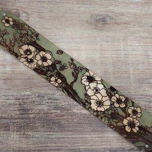 May include: A hand-tooled leather belt with a floral design. The belt features a light green background with a detailed illustration of white flowers and brown branches. The belt is secured with metal rivets.