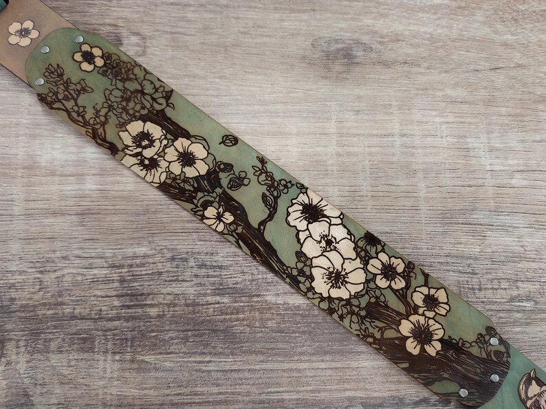 May include: A hand-tooled leather belt with a floral design. The belt features a light green background with a detailed illustration of white flowers and brown branches. The belt is secured with metal rivets.