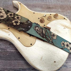 May include: A guitar strap with a teal and brown floral and bird design. The strap is made of leather and features a detailed illustration of a bird perched on a branch with white flowers. The strap is laid across a vintage white guitar.