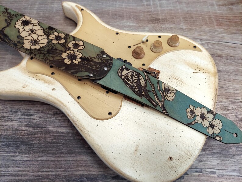 May include: A guitar strap with a teal and brown floral and bird design. The strap is made of leather and features a detailed illustration of a bird perched on a branch with white flowers. The strap is laid across a vintage white guitar.