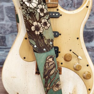 May include: A guitar strap with a nature-inspired design. The strap features a teal background with a detailed illustration of a bird perched on a branch, alongside a floral pattern. The strap is attached to a cream-colored electric guitar.