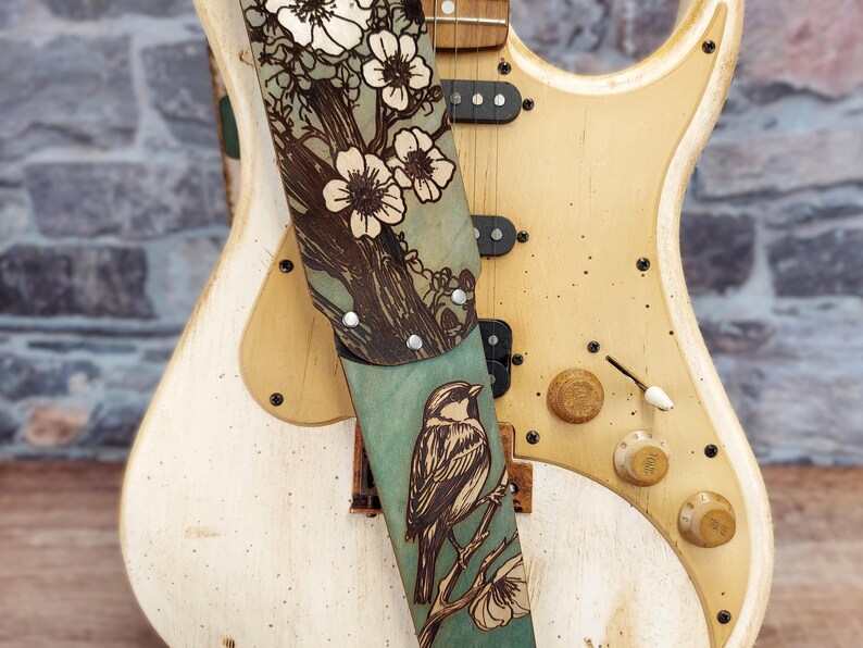 May include: A guitar strap with a nature-inspired design. The strap features a teal background with a detailed illustration of a bird perched on a branch, alongside a floral pattern. The strap is attached to a cream-colored electric guitar.