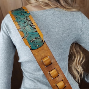 Puede incluir: Una correa de guitarra de cuero marr&oacute;n con un dise&ntilde;o detallado. La secci&oacute;n superior presenta una ilustraci&oacute;n de ola turquesa sobre un fondo amarillo. La correa tiene un patr&oacute;n repetitivo y tres lazos de cuero marr&oacute;n.