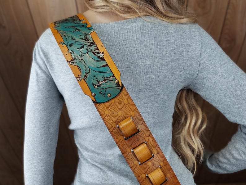 Puede incluir: Una correa de guitarra de cuero marr&oacute;n con un dise&ntilde;o detallado. La secci&oacute;n superior presenta una ilustraci&oacute;n de ola turquesa sobre un fondo amarillo. La correa tiene un patr&oacute;n repetitivo y tres lazos de cuero marr&oacute;n.