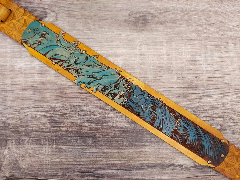 Puede incluir: Una correa de guitarra con un dise&ntilde;o detallado de olas. La correa es de cuero marr&oacute;n con un panel central que presenta una ilustraci&oacute;n de olas azules y negras. Los extremos tienen un patr&oacute;n decorativo y remaches met&aacute;licos.