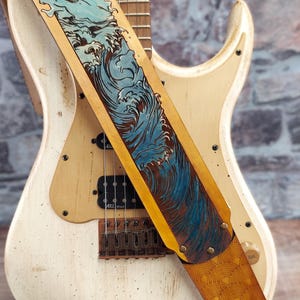 Puede incluir: Una guitarra el&eacute;ctrica crema con un acabado envejecido, con una correa de cuero. La correa es de color marr&oacute;n claro con un dise&ntilde;o de olas azules y un borde amarillo. La guitarra tiene pastillas negras y un puente marr&oacute;n.