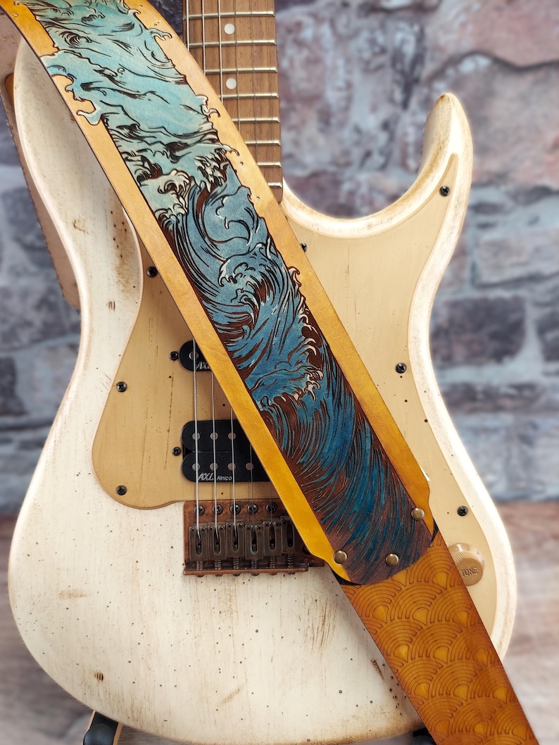 Puede incluir: Una guitarra el&eacute;ctrica crema con un acabado envejecido, con una correa de cuero. La correa es de color marr&oacute;n claro con un dise&ntilde;o de olas azules y un borde amarillo. La guitarra tiene pastillas negras y un puente marr&oacute;n.